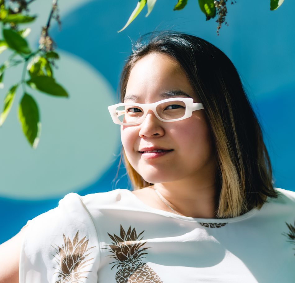 East Asian femme-presenting writer looking at camera and smiling with shoulder length brown hair with blonde tips and white contemporary-style glasses, wearing a white shirt with large gold pineapple print on a sky blue background.
