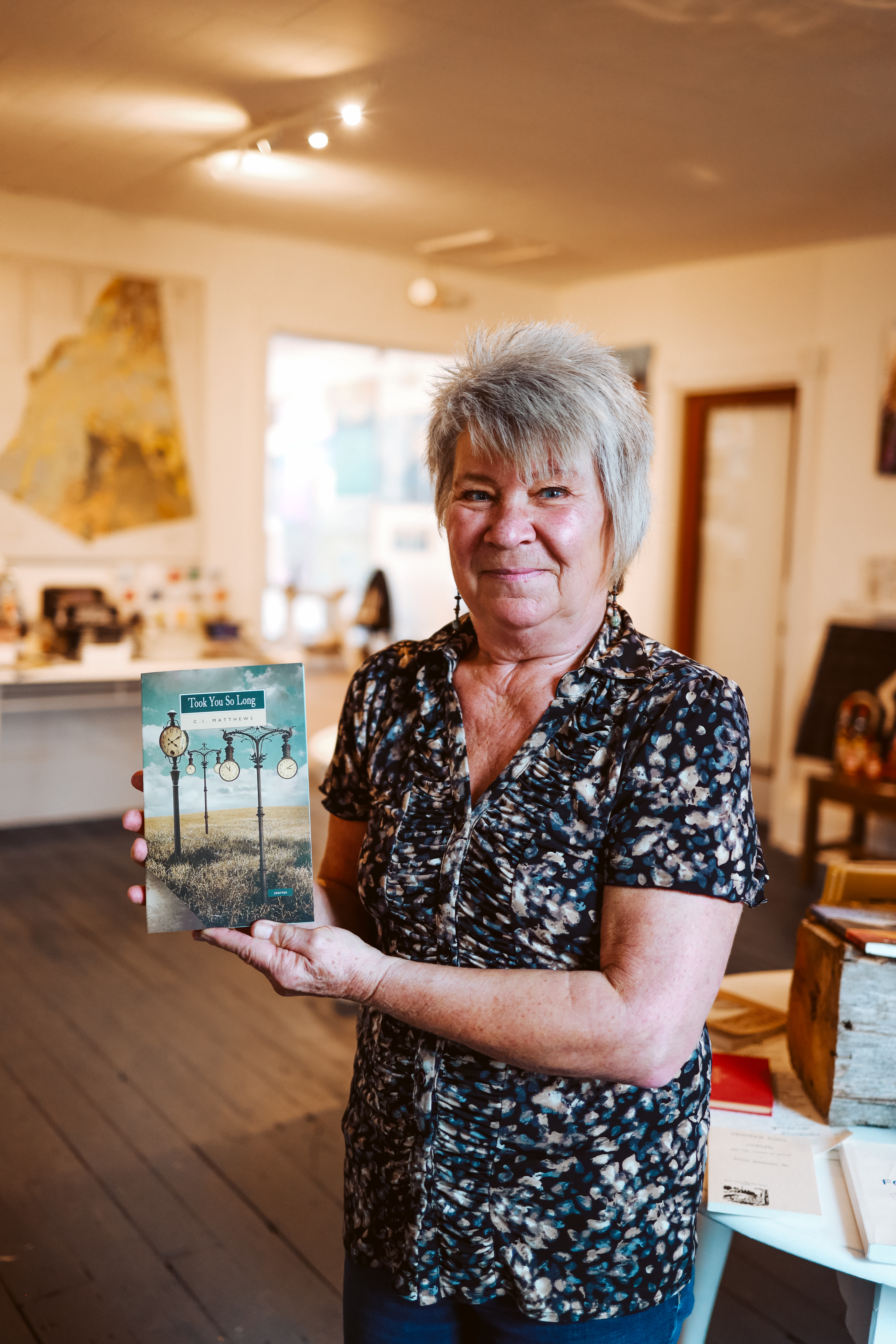 Author C. I. Matthews holding a copy of her award-winning short story collection Took You So Long. The background is the Mill Pond Gallery, Cargill, Ontario.
