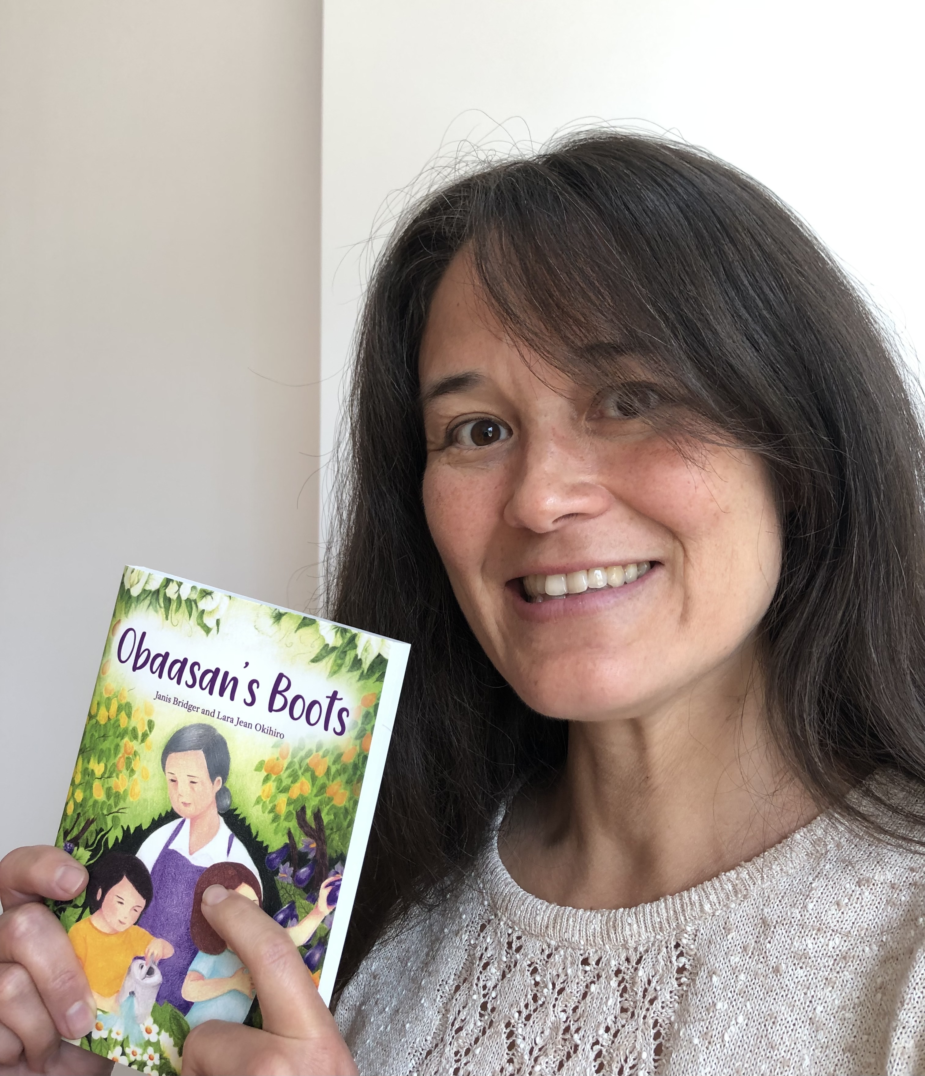 A woman with brown hair smiles holding a book that reads Obaasan's Boots.